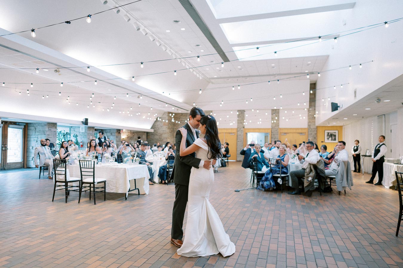 A bride and groom share their first dance at a wedding reception, surrounded by guests in a beautifully decorated venue with string lights.