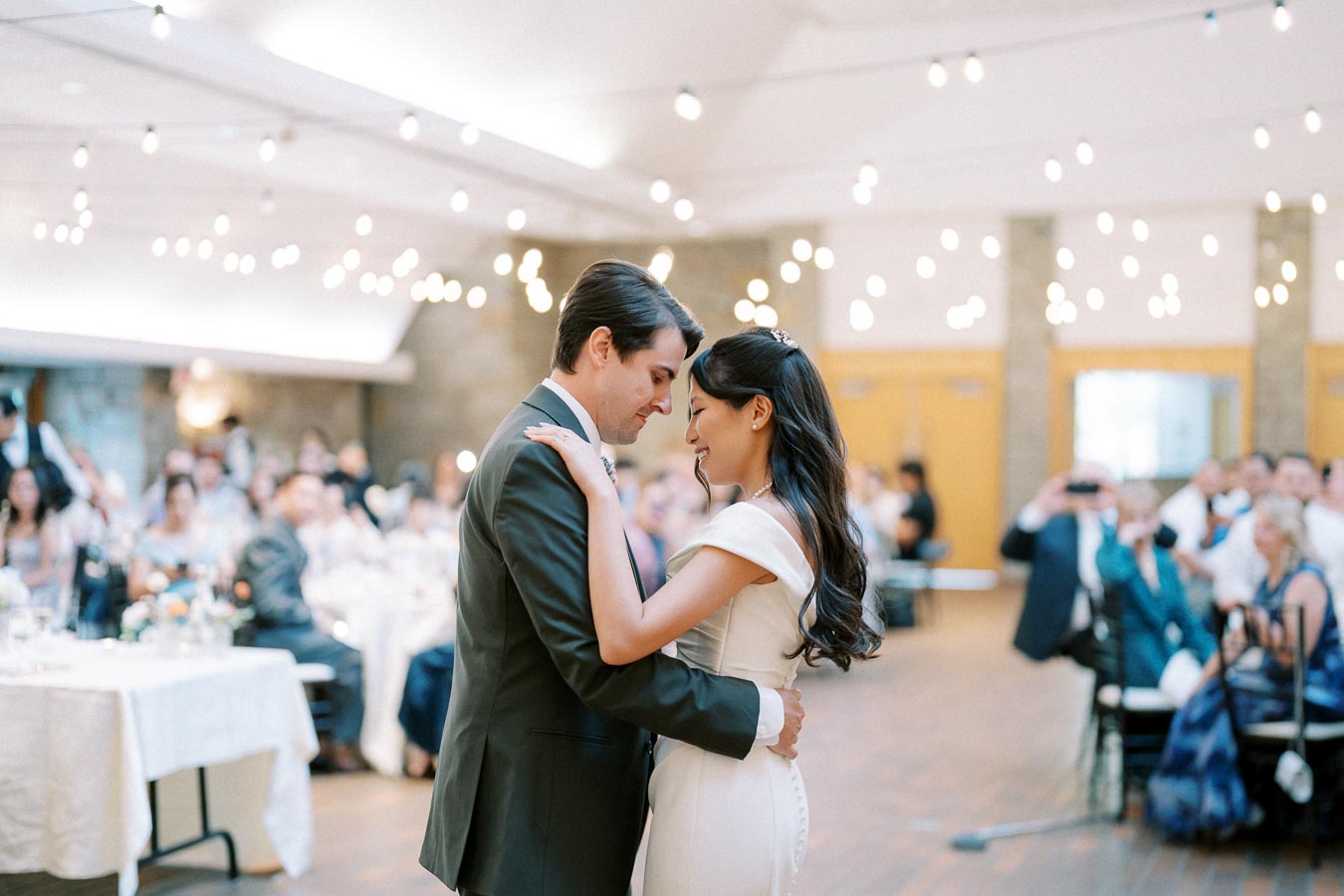 A newlywed couple shares a first dance at their wedding reception, surrounded by elegantly dressed guests and ambient string lights in a spacious venue.