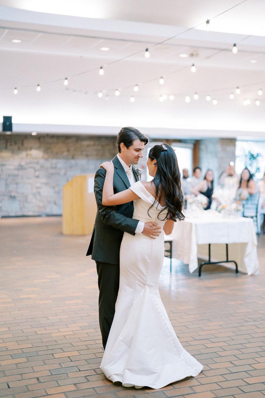A bride and groom share their first dance at a wedding reception, with overhead string lights creating a romantic ambiance and guests seated in the background.