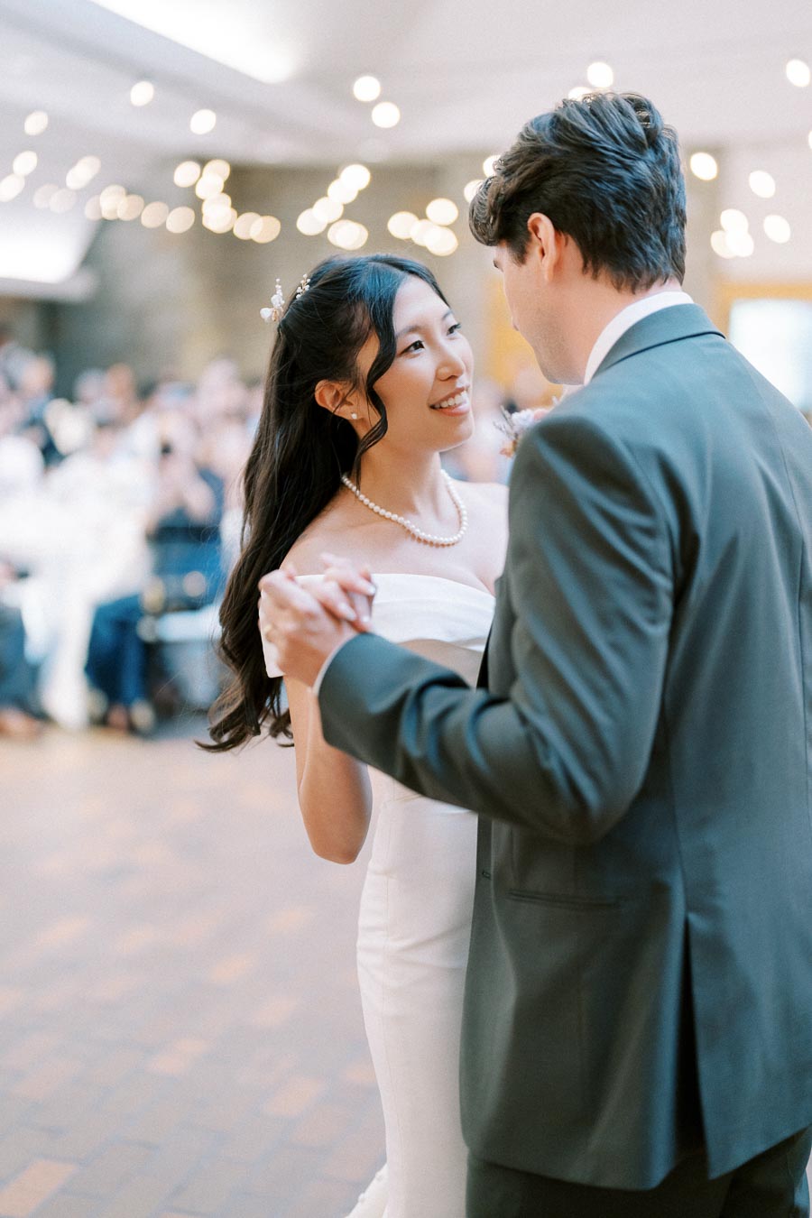A newlywed couple shares their first dance at a wedding reception, surrounded by softly glowing lights and a warmly lit room, with the bride wearing an elegant white dress and the groom in a classic suit.