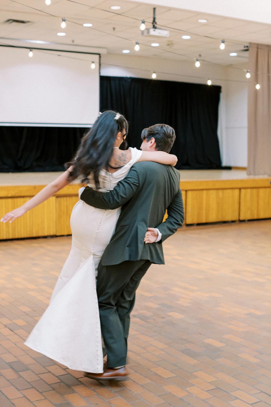 A couple elegantly dancing in an indoor event space, with the woman in a white dress and the man in a dark suit. The background features black curtains and string lights, creating a festive atmosphere.