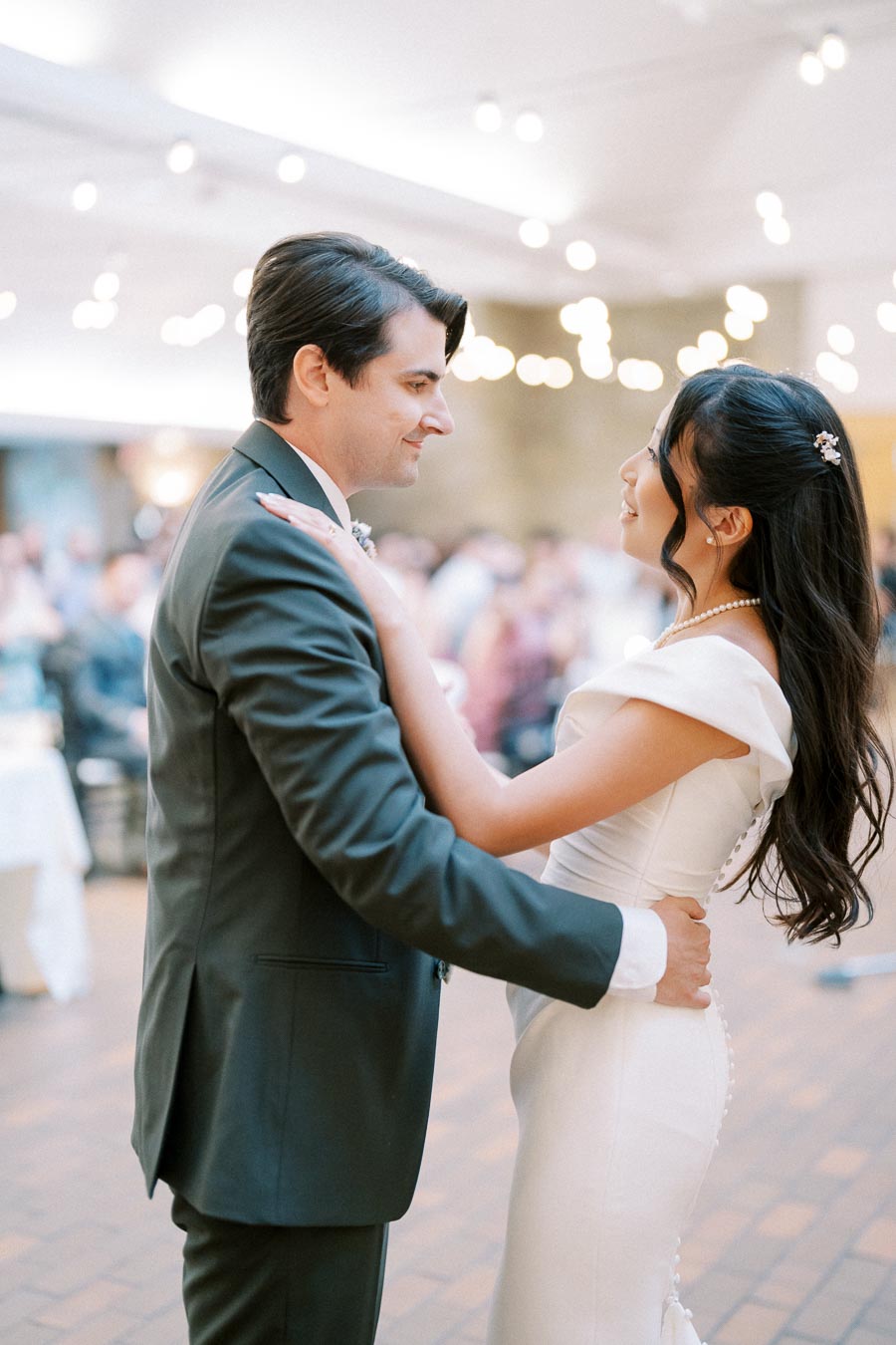 A bride and groom share their first dance at a wedding reception, surrounded by softly lit string lights, creating a romantic atmosphere.
