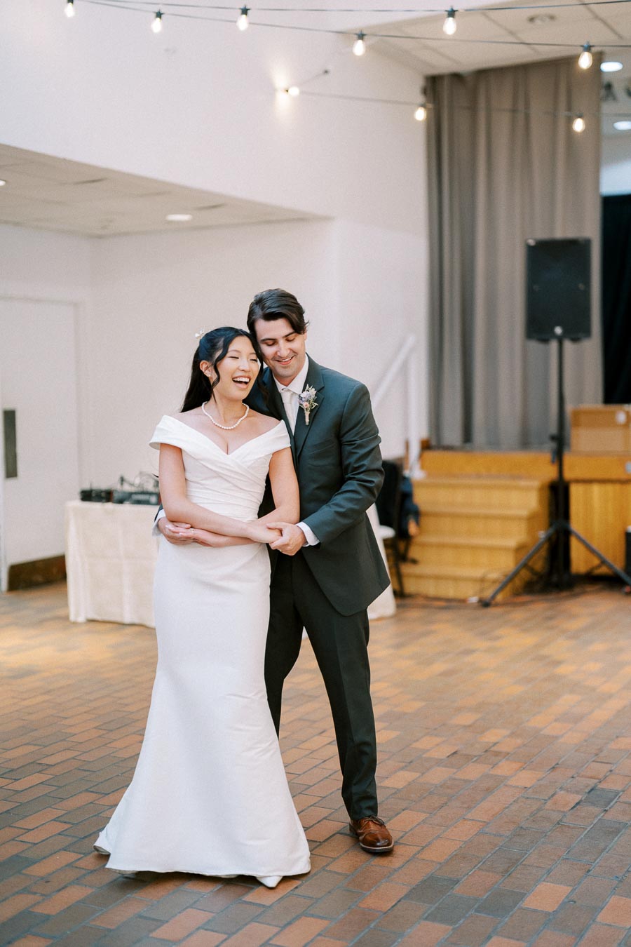 A joyful couple smiling during their wedding dance in an elegantly decorated venue, with string lights overhead and a dance floor beneath them.