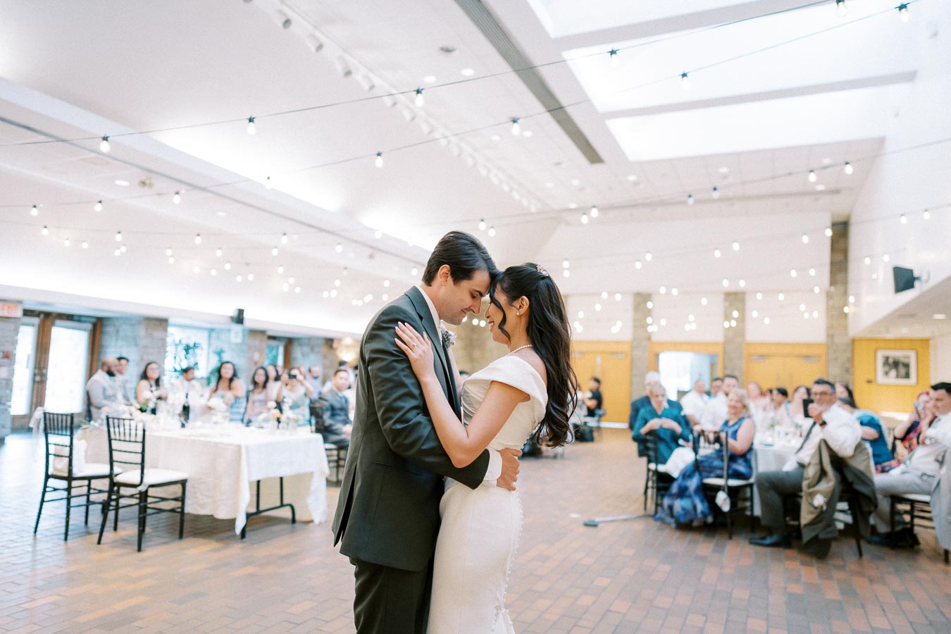 Couple shares a romantic first dance at a wedding reception, surrounded by guests in an elegantly decorated venue with string lights.