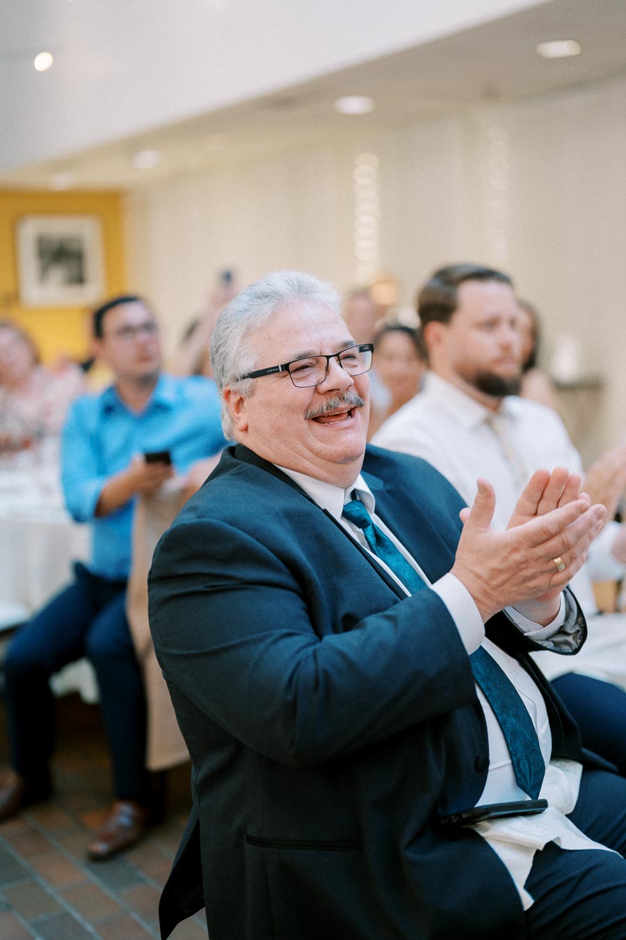 A man in a suit and tie, glasses, and a mustache applauding joyfully at an indoor event, surrounded by a blurred audience.