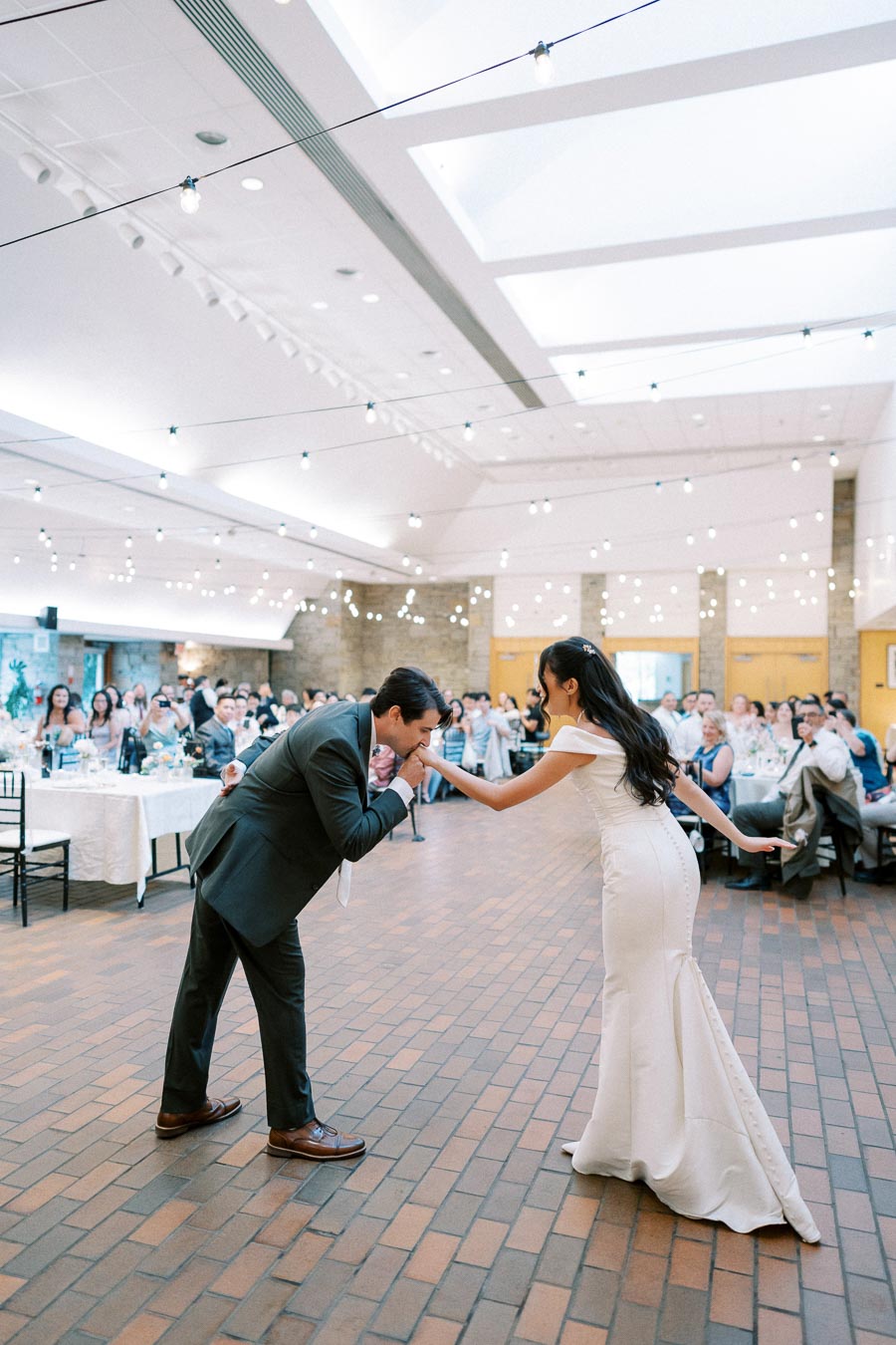 A bride in a white dress and a groom in a suit share a romantic moment at their wedding reception in a decorated venue with string lights and guests seated at tables in the background.