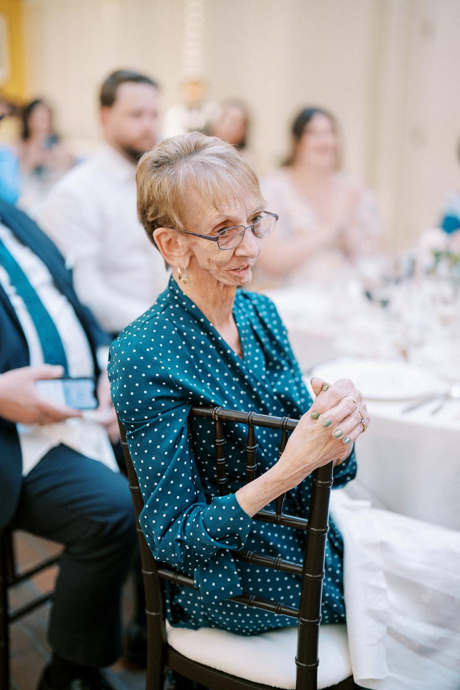 Elderly woman in a polka dot dress smiling while seated at a formal event, surrounded by guests in a banquet setting.