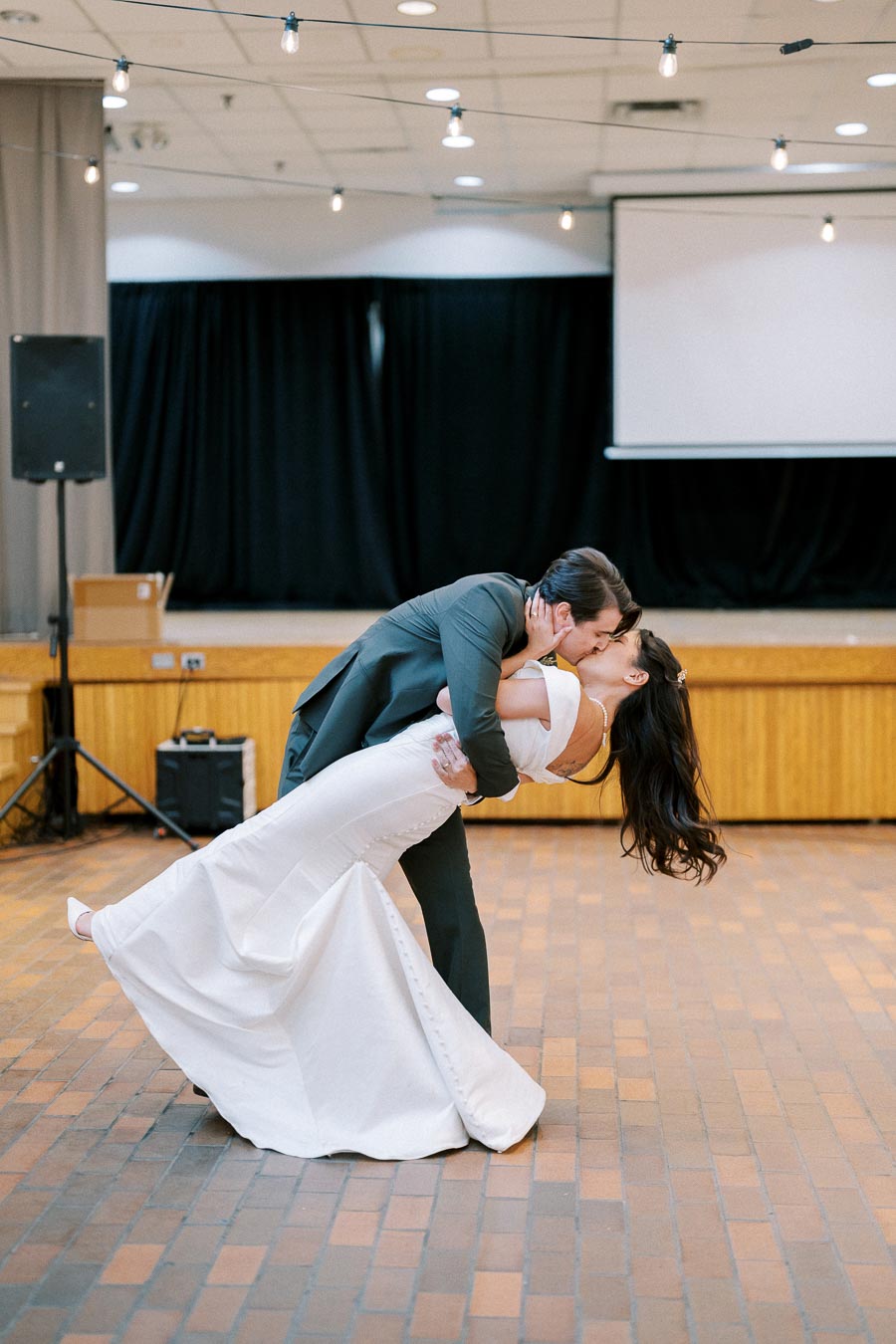 A couple sharing a romantic dance at a wedding, with the groom dipping the bride while kissing her in a warmly lit reception hall. The bride is wearing an elegant white gown, and the groom is in a dark suit.