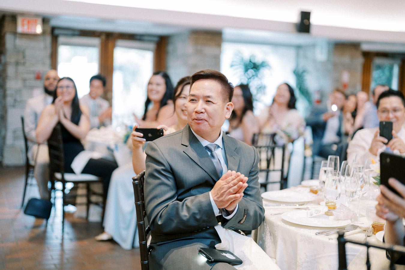 A man in a suit clapping at a wedding reception, surrounded by seated guests who are smiling and taking photos in a warmly lit venue.
