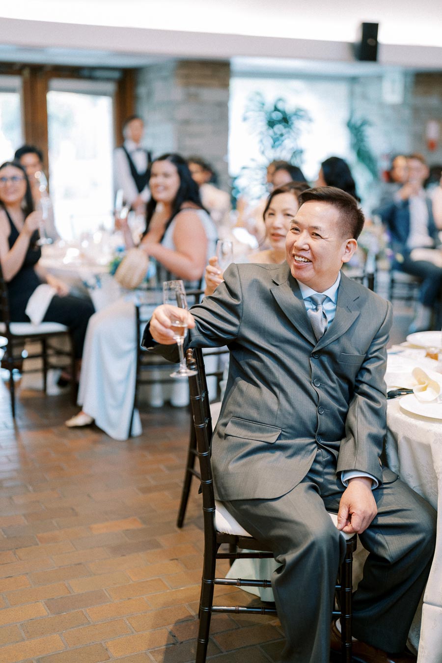 A smiling man in a suit holding a champagne glass at a celebratory event, with guests seated at tables in the background.