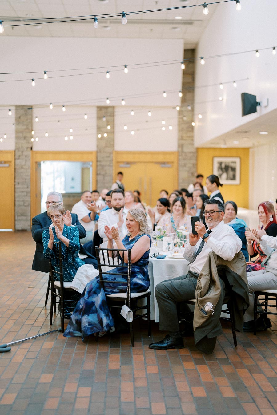 A joyful wedding reception inside a warmly lit venue with guests clapping and smiling at round tables, capturing the festive atmosphere of the celebration.