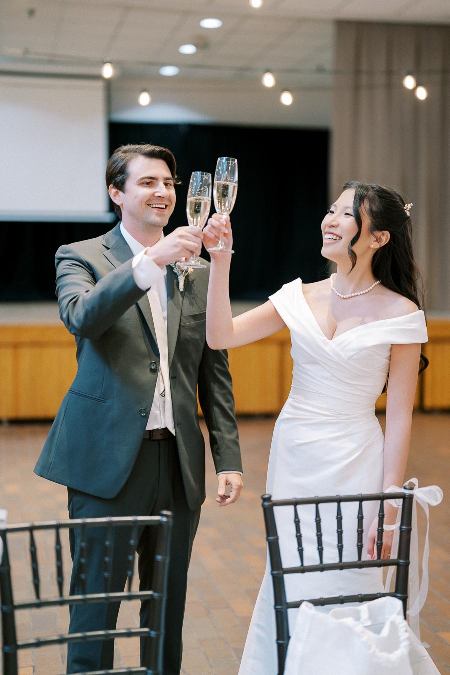 Two people in elegant wedding attire raising champagne glasses in celebration, smiling joyfully at a formal event with chairs and lights in the background.