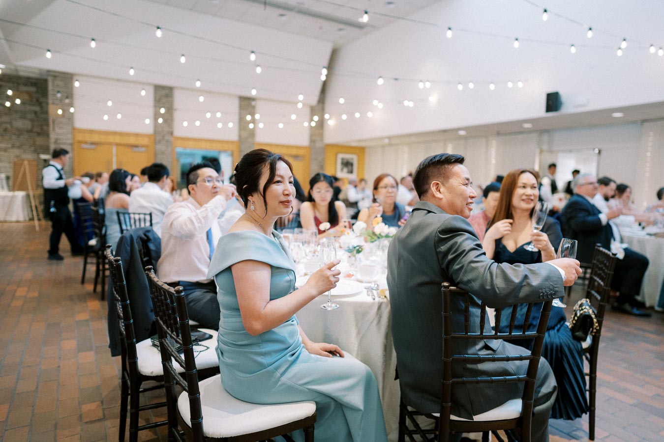 Guests seated at elegantly decorated tables during a wedding reception, with overhead string lights creating a festive atmosphere. Everyone is dressed formally, enjoying drinks and socializing in a bright and spacious venue.