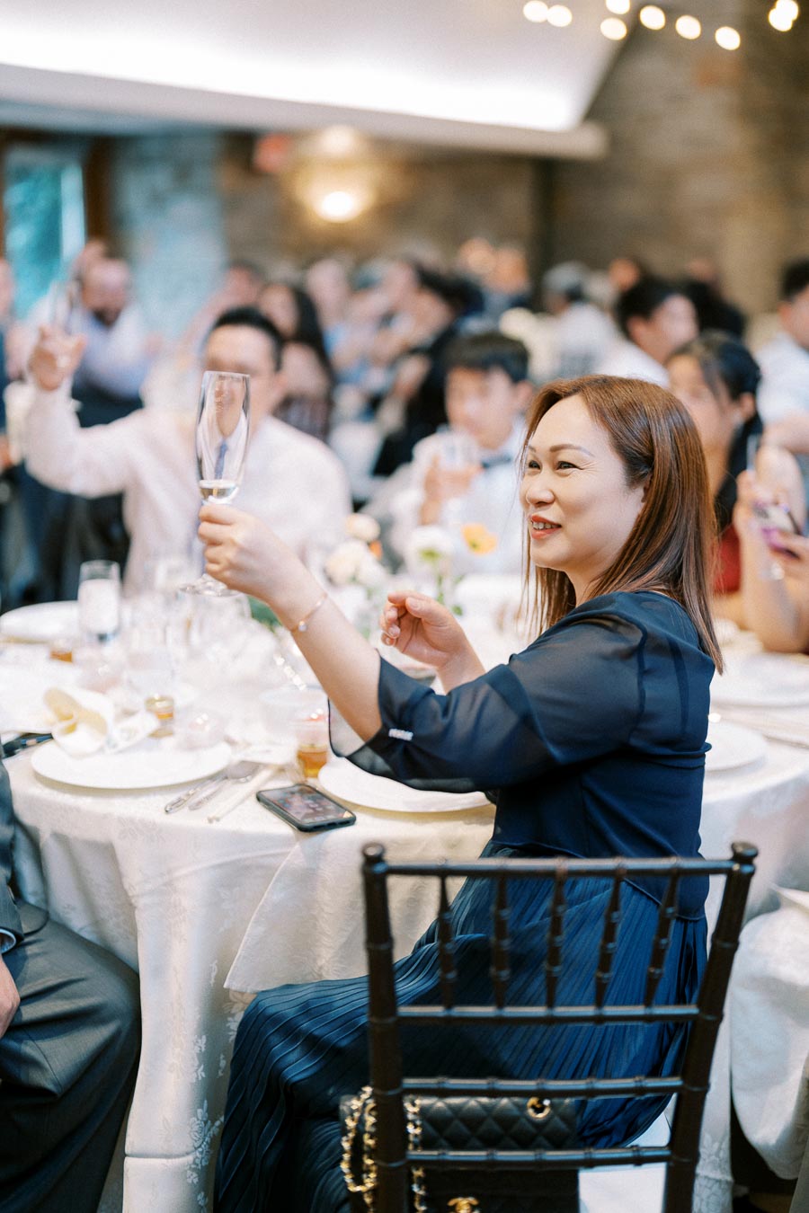 A woman in a blue dress raises a toast at a formal event, surrounded by elegantly set tables and a large group of guests celebrating in an atmospheric venue.