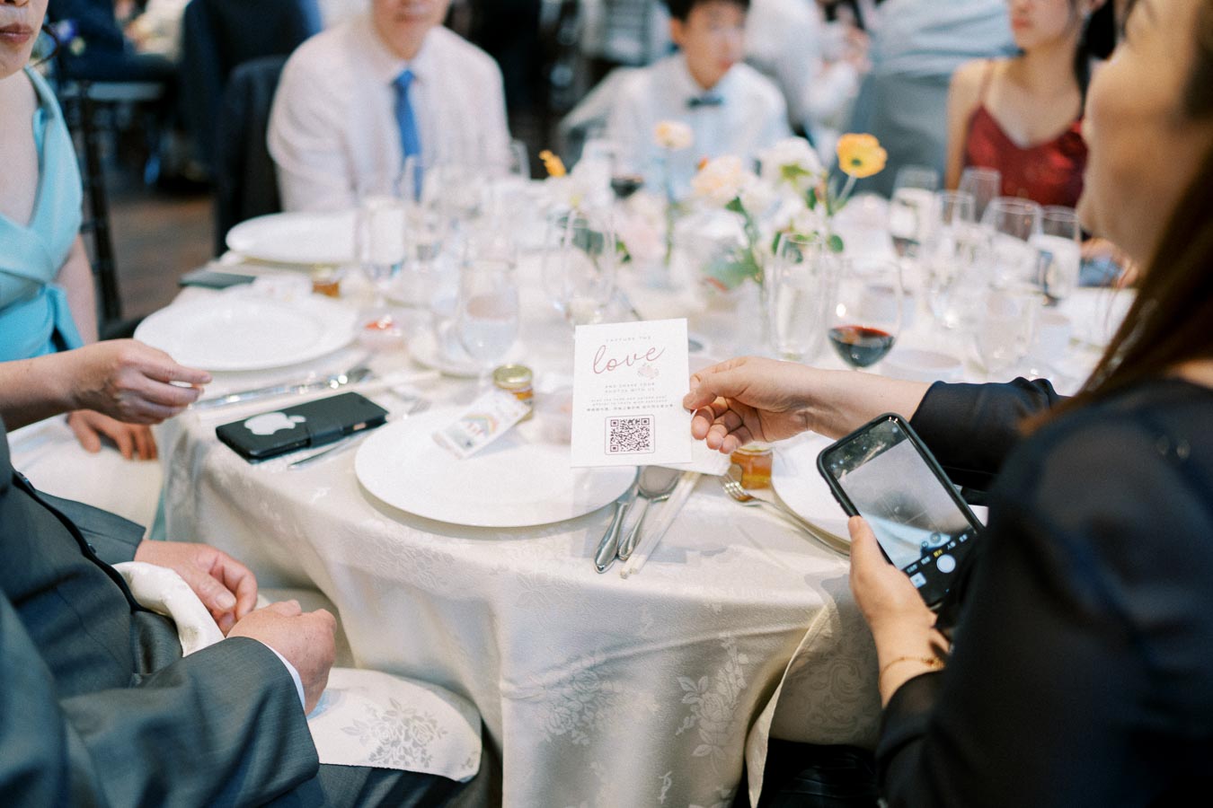 A group of elegantly dressed people sitting around a wedding reception table with a white tablecloth, holding a card with love and a QR code, as one person uses a smartphone to scan it. Plates, glassware, and floral centerpieces decorate the table.