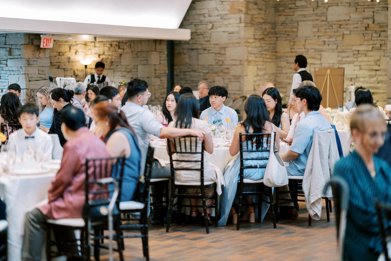 Guests seated at elegantly decorated tables in a rustic venue, engaged in conversation during a formal event or celebration, with soft lighting and a stone wall background.