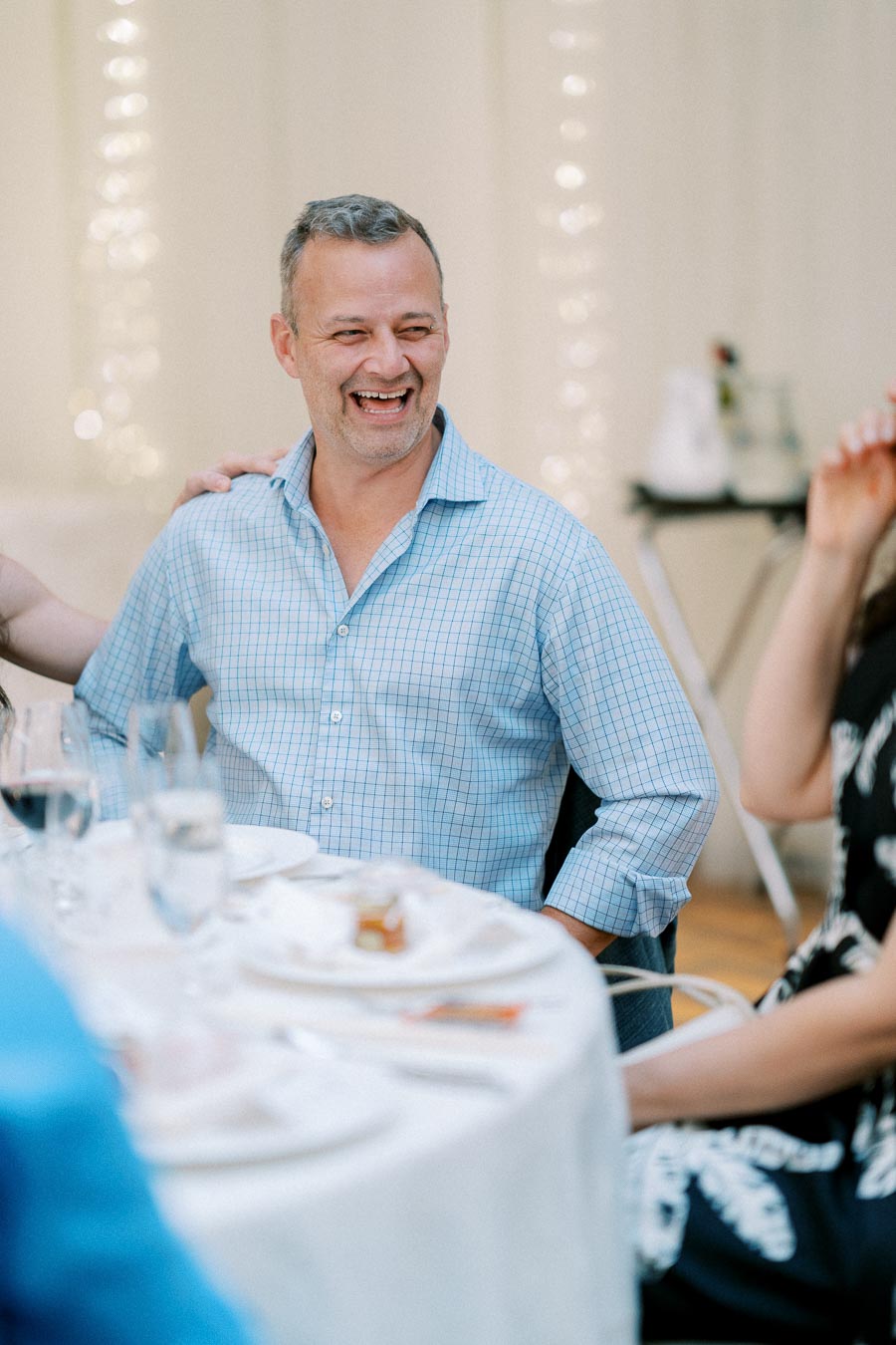 A man in a blue checkered shirt laughing at a dining table during a gathering, with plates and wine glasses in the foreground and soft lighting in the background.