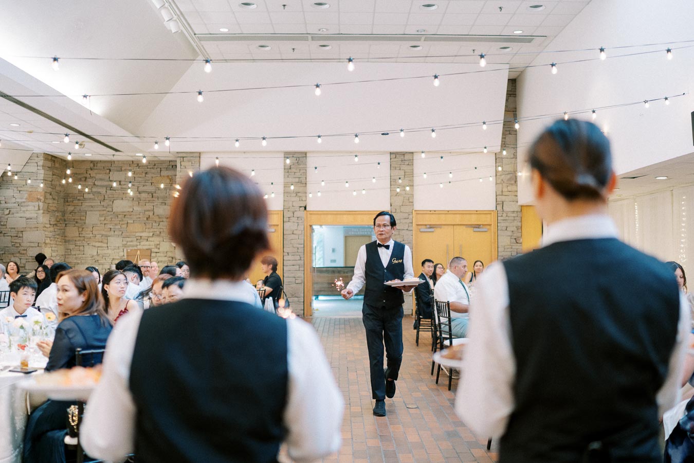 A waiter in a formal dining setting serving a crowded banquet hall, wearing a vest with a name tag, under string lights with a stone wall backdrop.