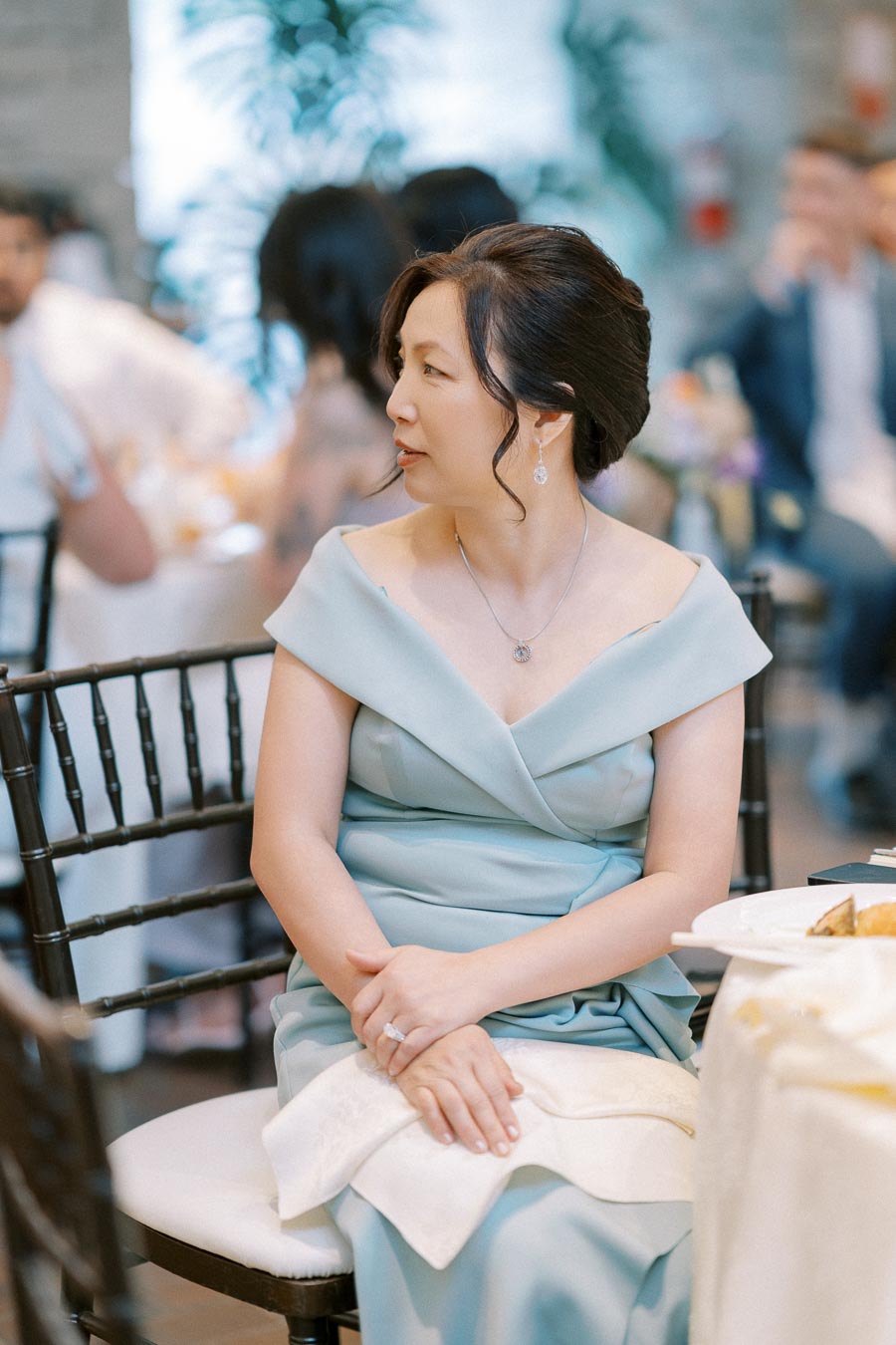 A woman in an elegant light blue dress sits at a wedding reception, looking sideways. She has an updo hairstyle and is wearing a necklace and earrings. The background features guests and soft lighting, creating a warm, inviting atmosphere.