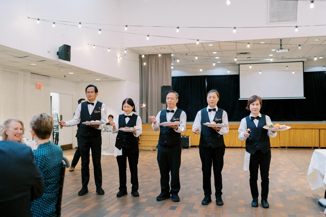 Five servers wearing matching uniforms hold sparklers and plates of food during an elegant restaurant event with warm lighting.