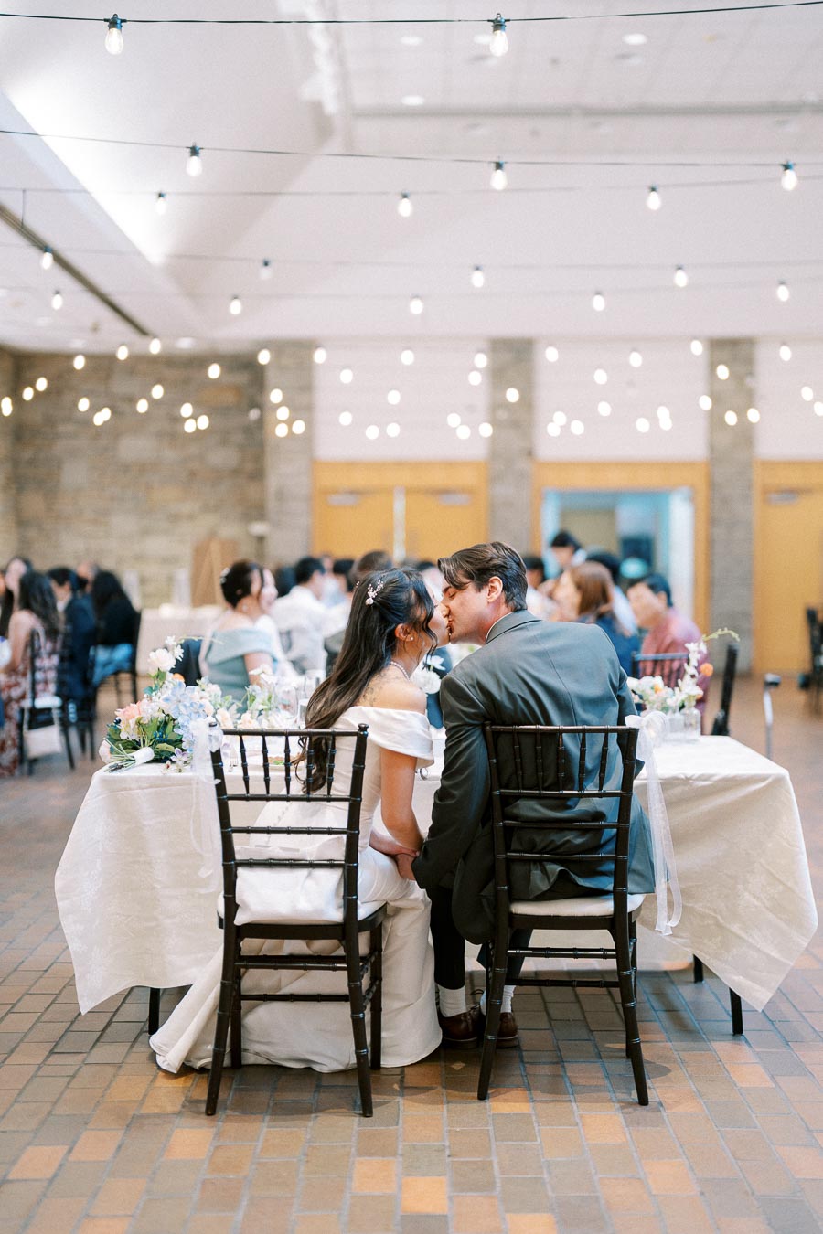 Elegant wedding reception with bride and groom sharing a romantic moment at a decorated table, surrounded by soft string lights and seated guests.