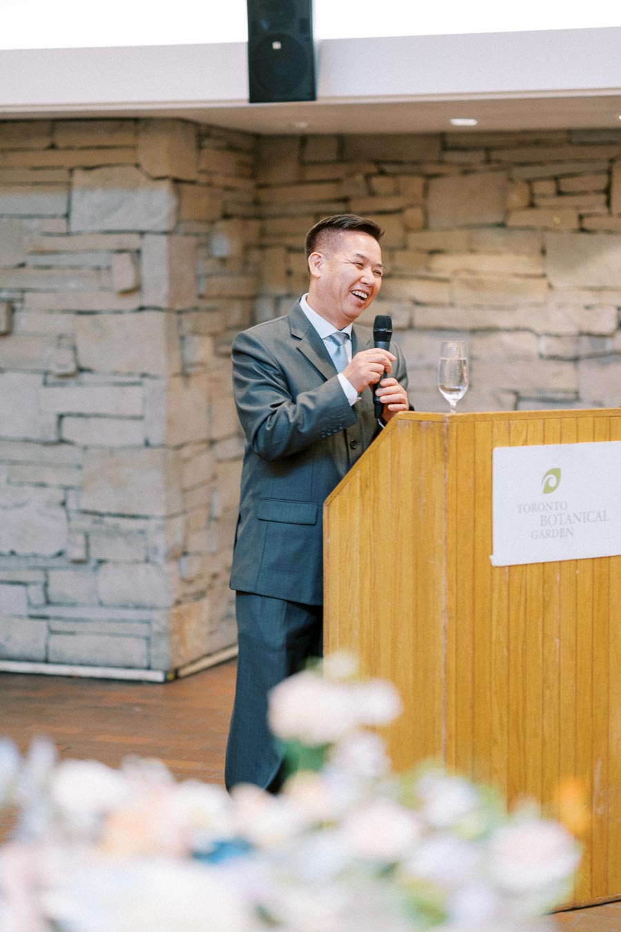 A man in a suit joyfully gives a speech at a podium in Toronto Botanical Garden, holding a microphone and smiling.