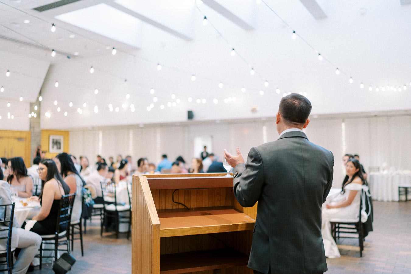 A man in a suit giving a speech at a wedding reception, standing behind a wooden podium. The room is elegantly decorated with string lights, and guests are seated at round tables, attentively listening. The atmosphere is festive and formal, suitable for a wedding celebration.