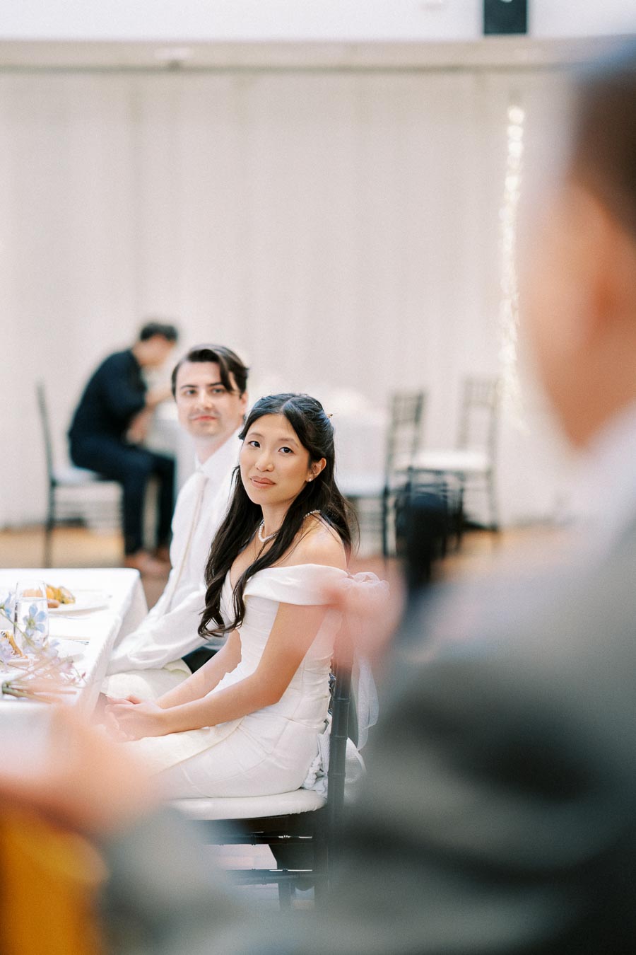Wedding reception with a bride and groom seated at a table, the bride in a white dress looking towards the camera with soft lighting and a blurred background.