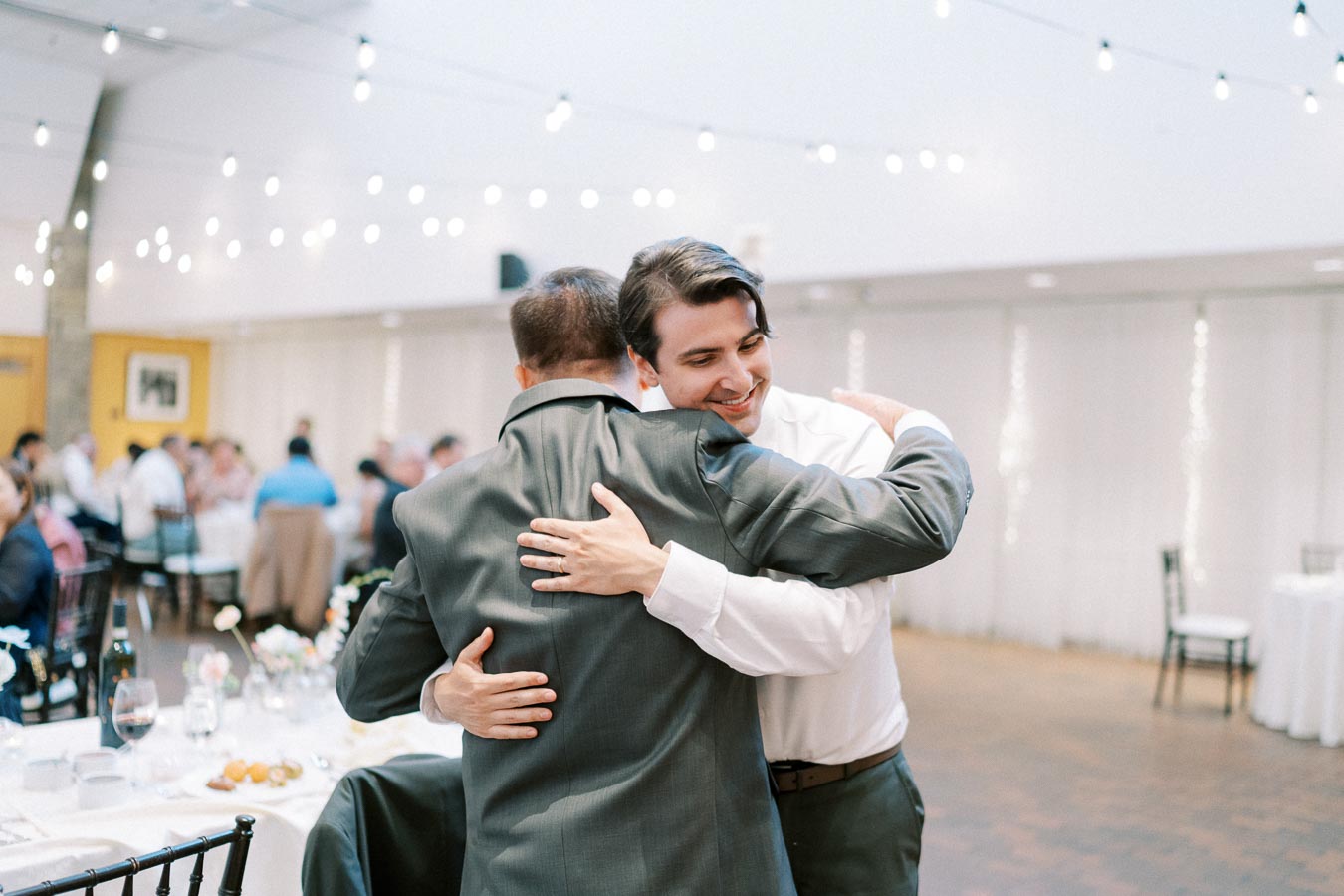Two men embracing warmly at an indoor event with tables and string lights in the background, conveying a joyful reunion.