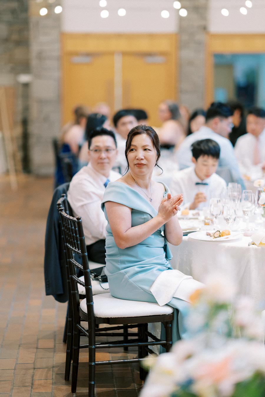 A woman in a blue dress applauding at a formal event, seated at a table with elegantly set dining arrangements, surrounded by other guests in a softly lit banquet hall.