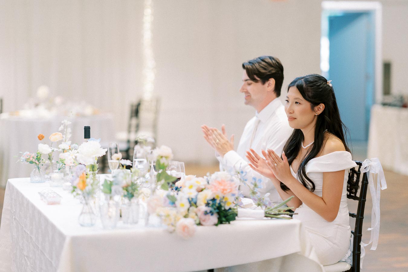 A bride and groom sitting at a beautifully decorated wedding reception table, adorned with pastel flowers and elegant glassware, both clapping and smiling during a celebration.