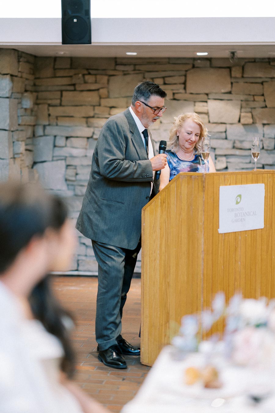 A man and woman deliver a speech at a podium inside a stone-walled venue during a formal event at Toronto Botanical Garden.