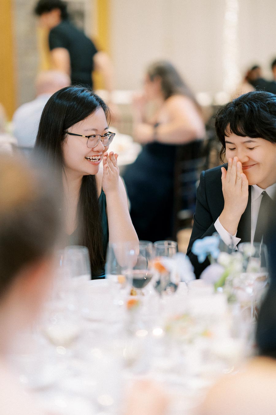 Two women sharing a light-hearted moment and smiling at a formal event, surrounded by elegantly set tables with flowers and glassware.