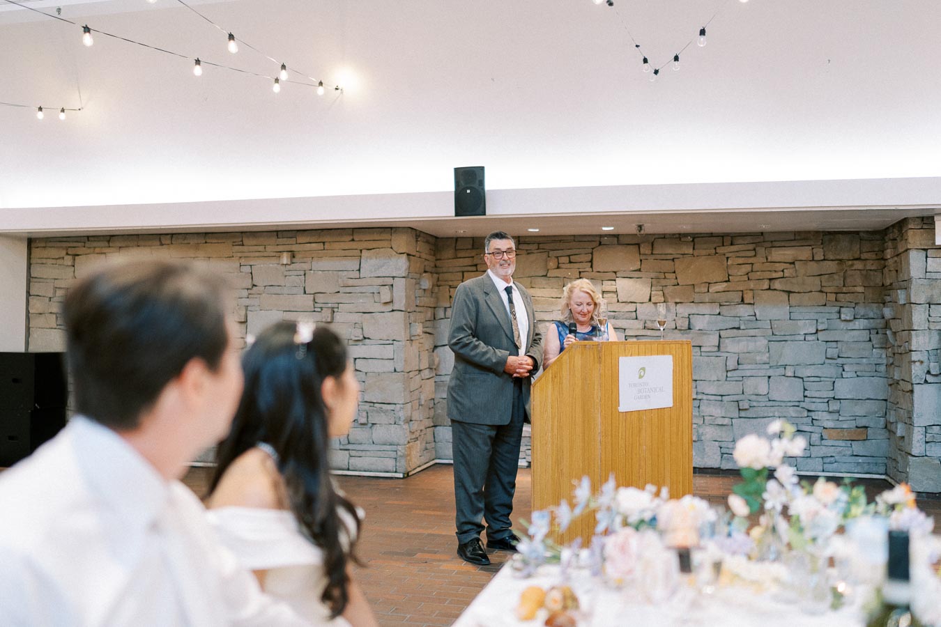 Wedding reception scene with a man in a suit and a woman at a podium giving a speech, while guests attentively listen at a table in the foreground adorned with flowers. The setting features a stone wall backdrop, creating a warm and intimate atmosphere.