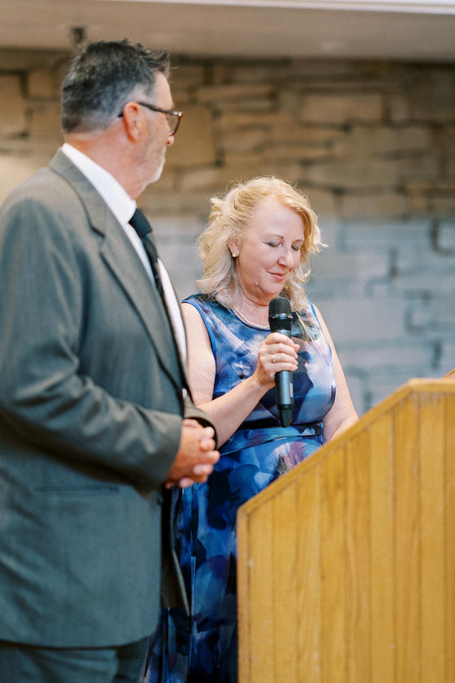 A woman in a blue dress holds a microphone while standing next to a man in a suit. They are at a podium inside a room with a stone wall background.