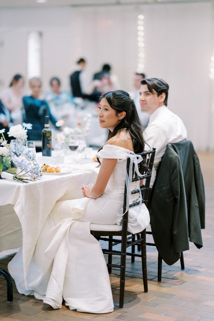 A bride and groom seated at a beautifully decorated wedding reception table, surrounded by guests in a bright venue.