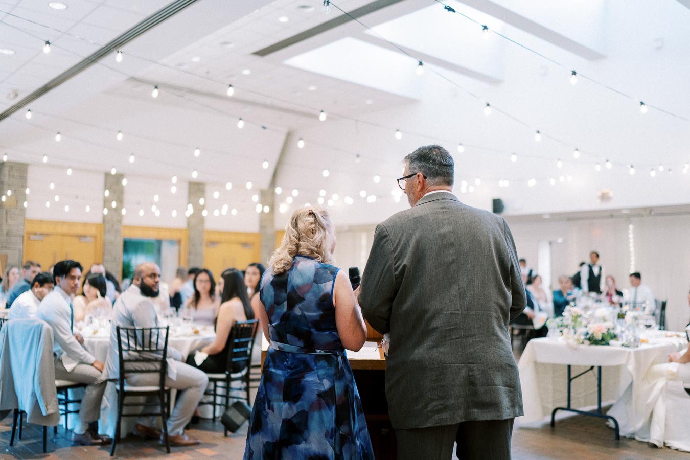 A couple giving a speech at a wedding reception, surrounded by seated guests in a warmly lit banquet hall with string lights and elegantly set tables.
