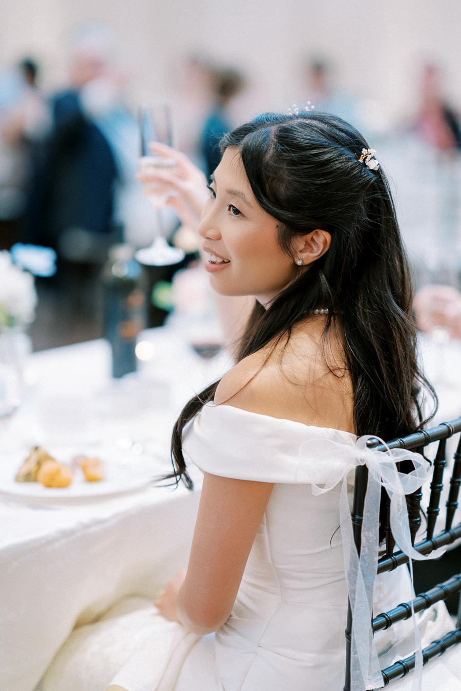 Young woman in an elegant off-shoulder white dress sitting at a wedding reception, holding a champagne glass with a smile, surrounded by a softly blurred background of attendees and table setting.