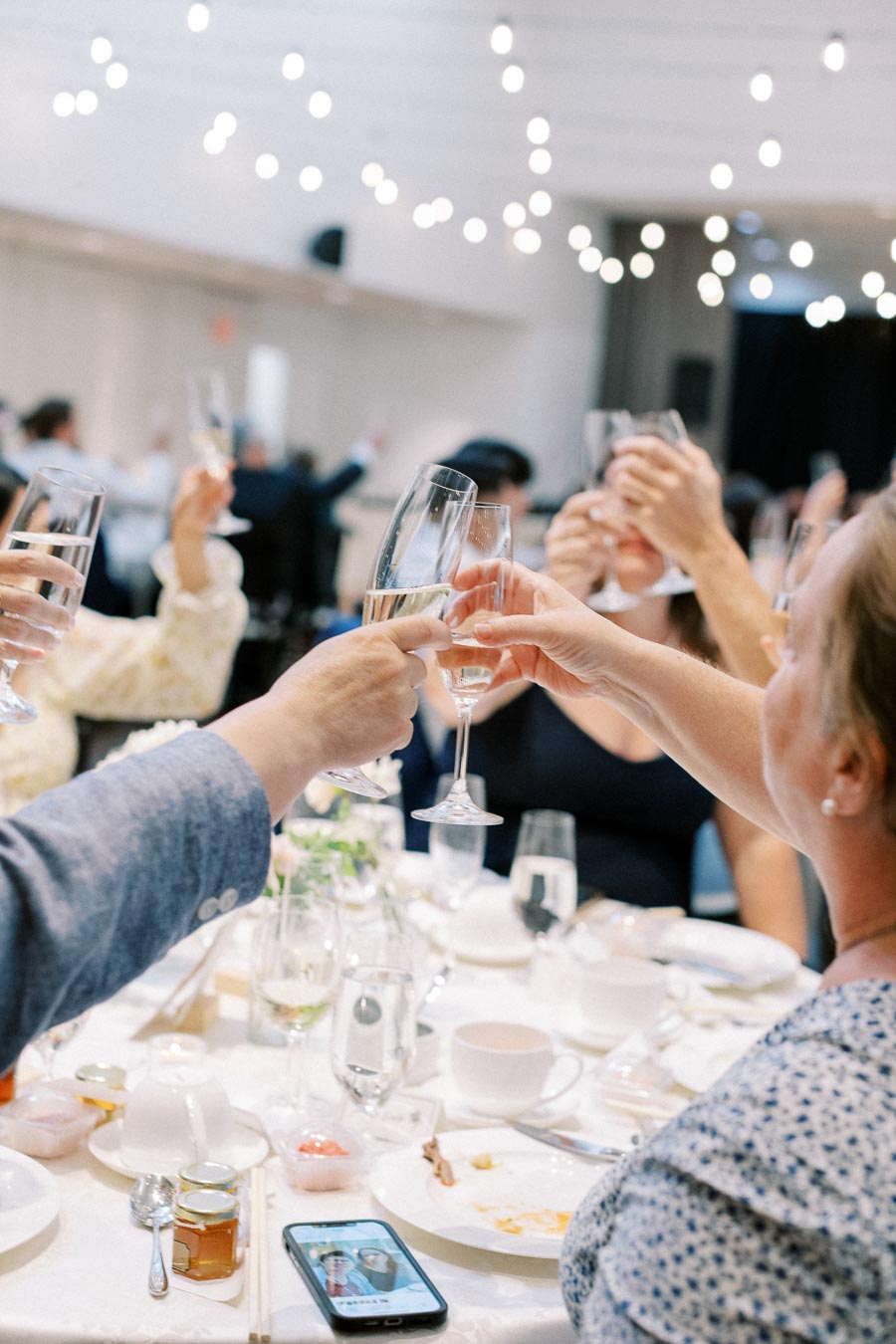 Group of people raising champagne glasses in a celebratory toast at an elegant indoor gathering, with a table set with dishes and decorations beneath twinkling string lights.