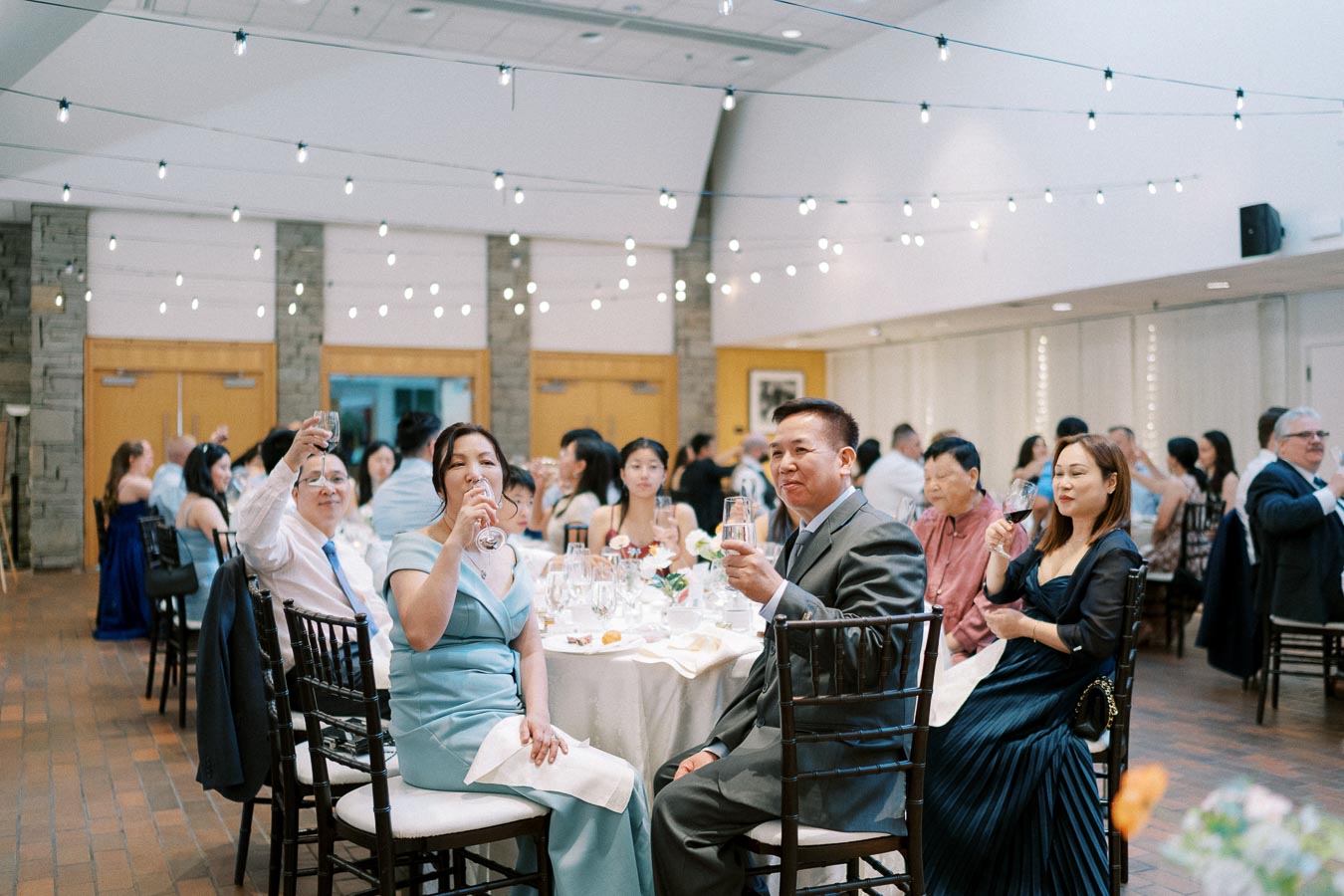 A group of elegantly dressed guests raising their glasses in a toast at a formal indoor wedding reception, adorned with string lights and set with round tables.