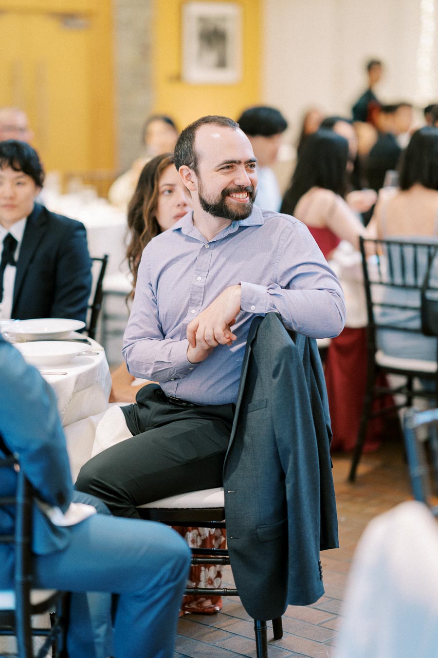A man in a formal shirt sits at a wedding reception table, smiling and interacting with other guests, with a jacket draped over the back of his chair.