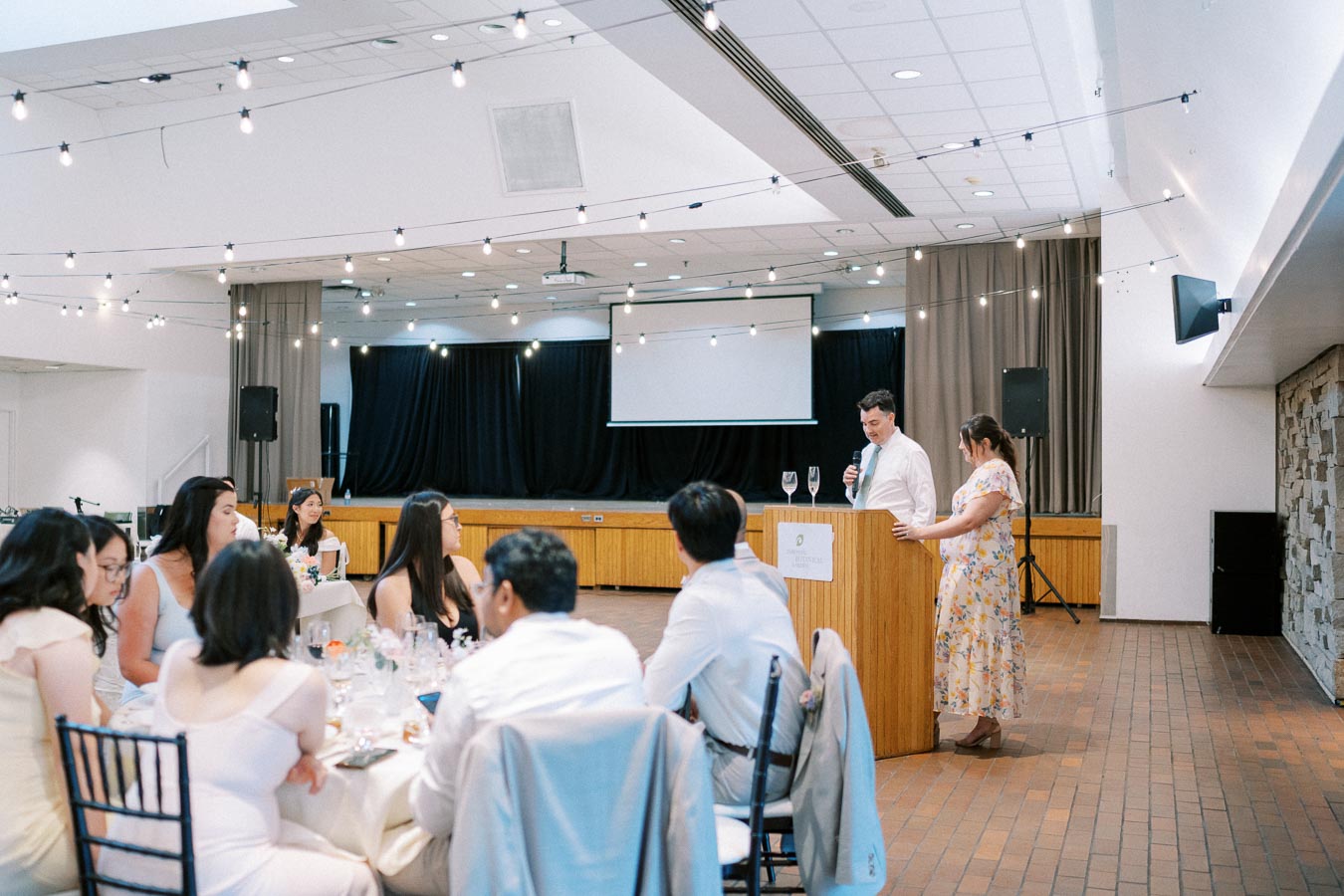 Wedding reception scene with guests seated at tables adorned with flowers, while a speaker gives a speech alongside a woman standing at a podium. The venue features elegant string lighting and a presentation screen in the background.