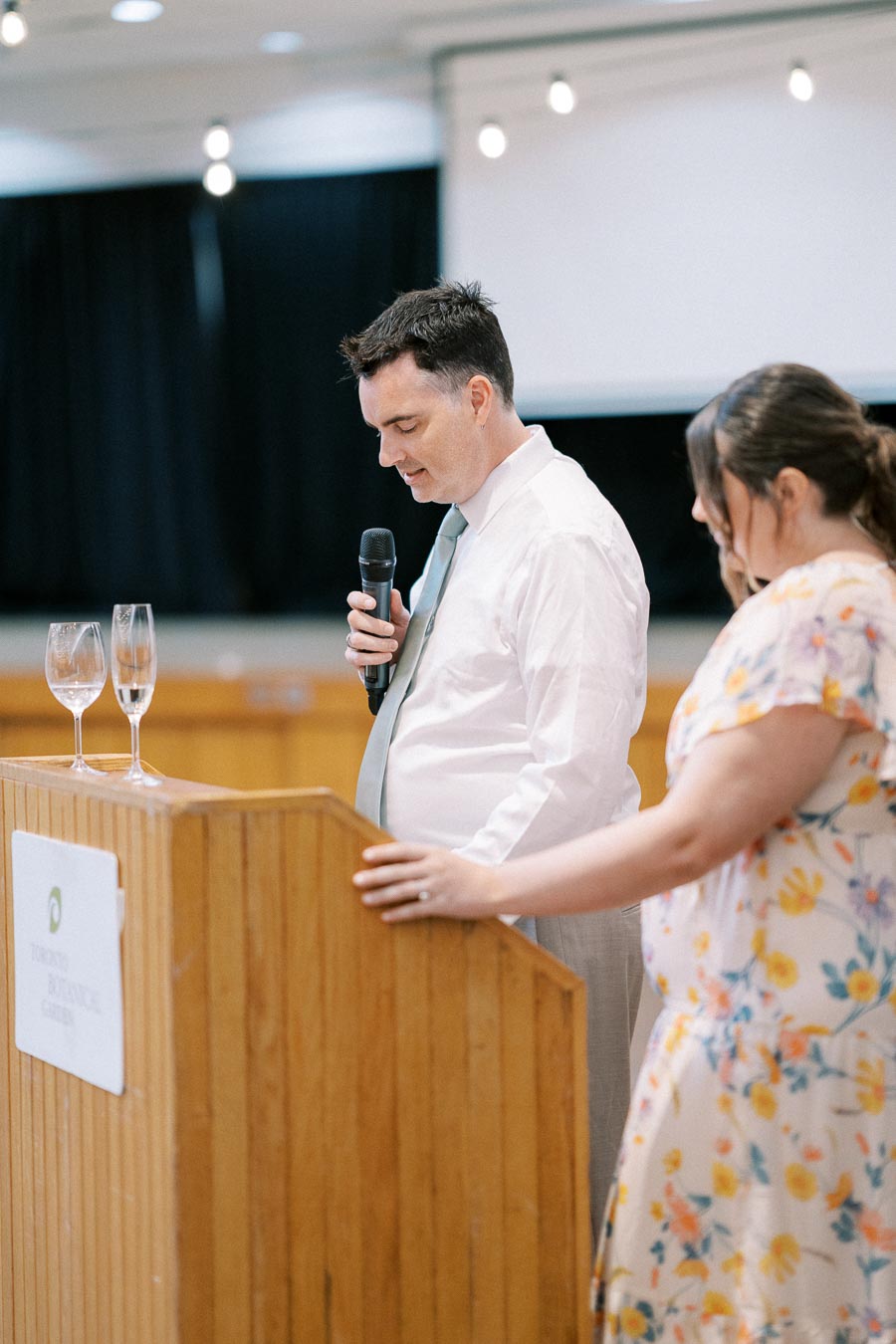 Man in a white shirt speaking into a microphone at a wooden podium, with a woman in a floral dress by his side, set in an indoor venue with dim lighting and wine glasses on the podium.