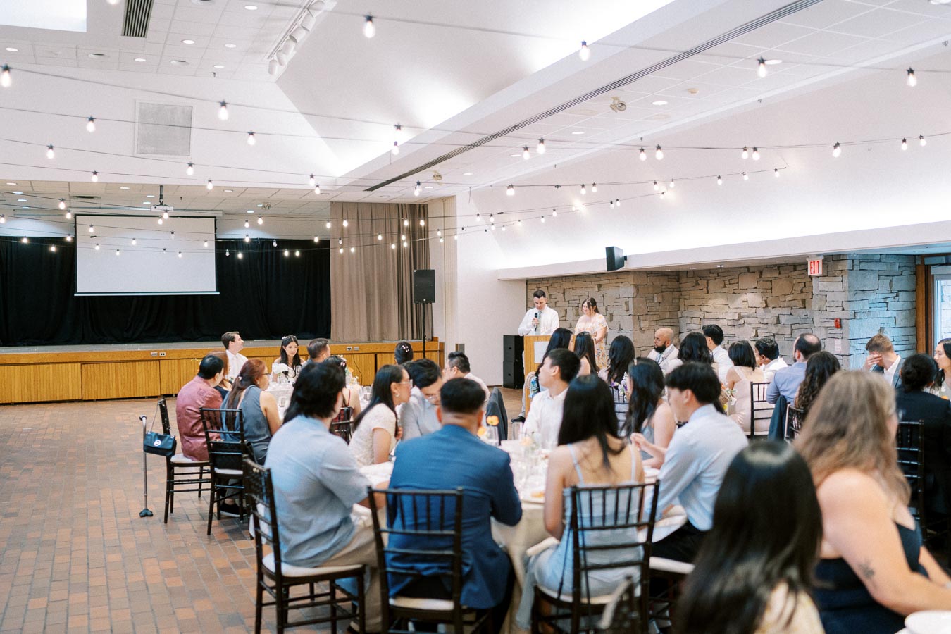 A well-decorated wedding reception venue with string lights hanging from the ceiling, guests seated around round tables, and a speaker at a podium in the background.