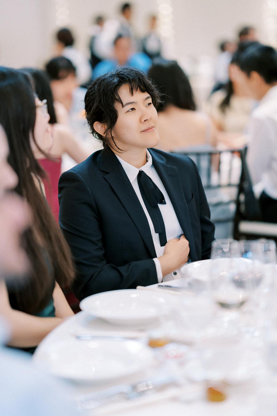 A person in formal attire sits attentively at a elegantly set dining table during an event, surrounded by other guests in a softly lit room.