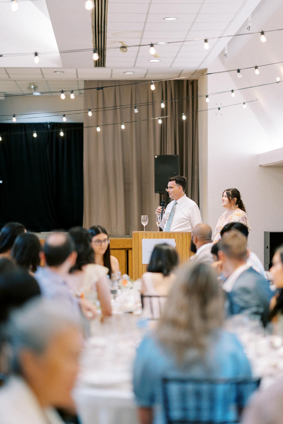Man giving a speech at a wedding reception, standing at a podium with a microphone, surrounded by guests seated at round tables, under string lights in a decorated hall.