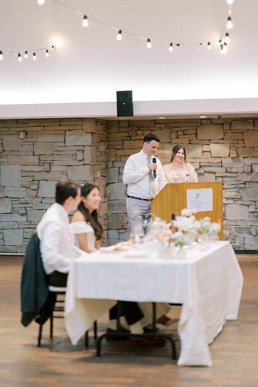 Wedding reception at Toronto Botanical Garden with a man giving a speech at a podium, while guests sit at a table adorned with flowers and a stone wall backdrop under string lights.