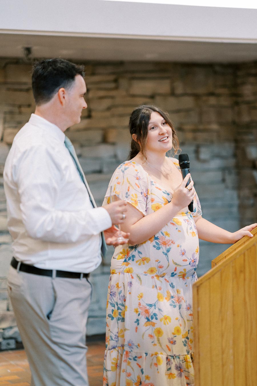 Two people speaking at an indoor event, one holding a microphone and wearing a floral dress, while the other in a white shirt looks on.