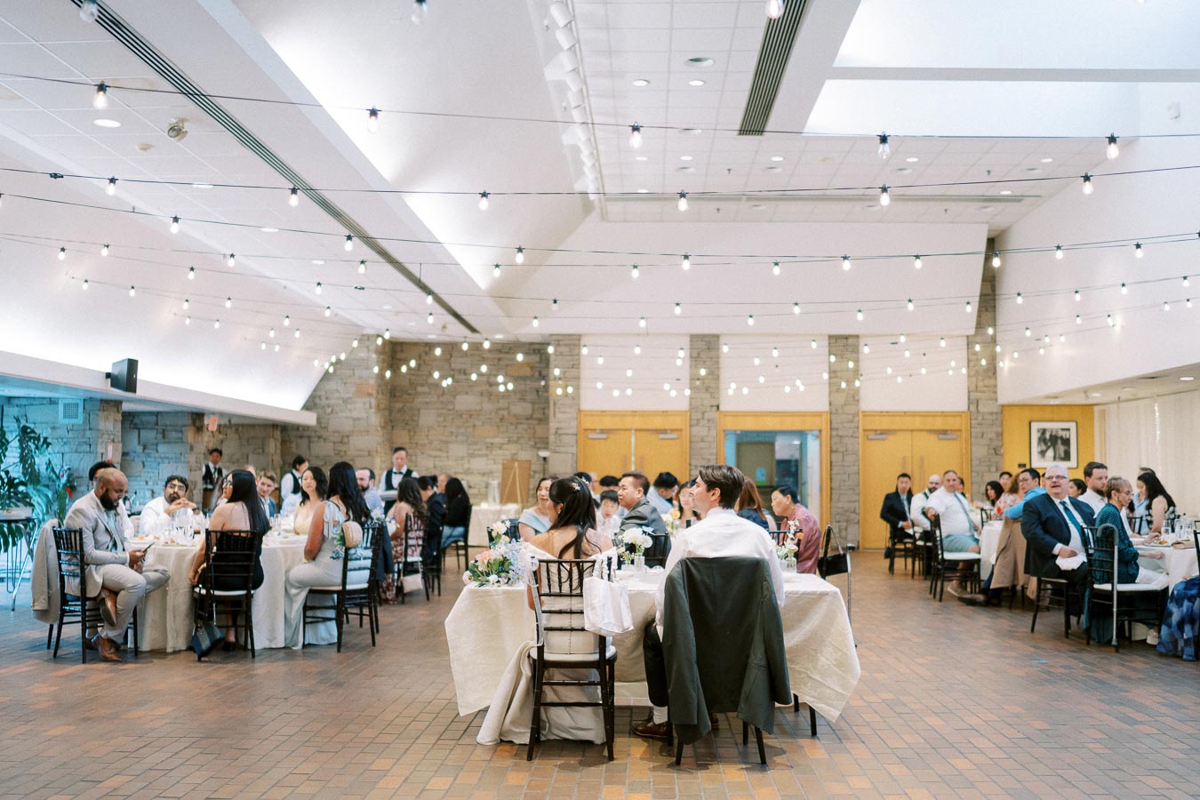 Wedding reception in a decorated banquet hall with guests seated at tables, ambient string lights overhead, and a stone wall backdrop.