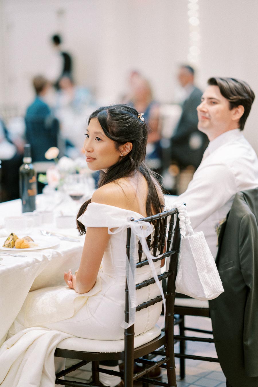 A bride in an elegant white gown sits at a decorated table during a wedding reception, with a blurred background of guests in formal attire.